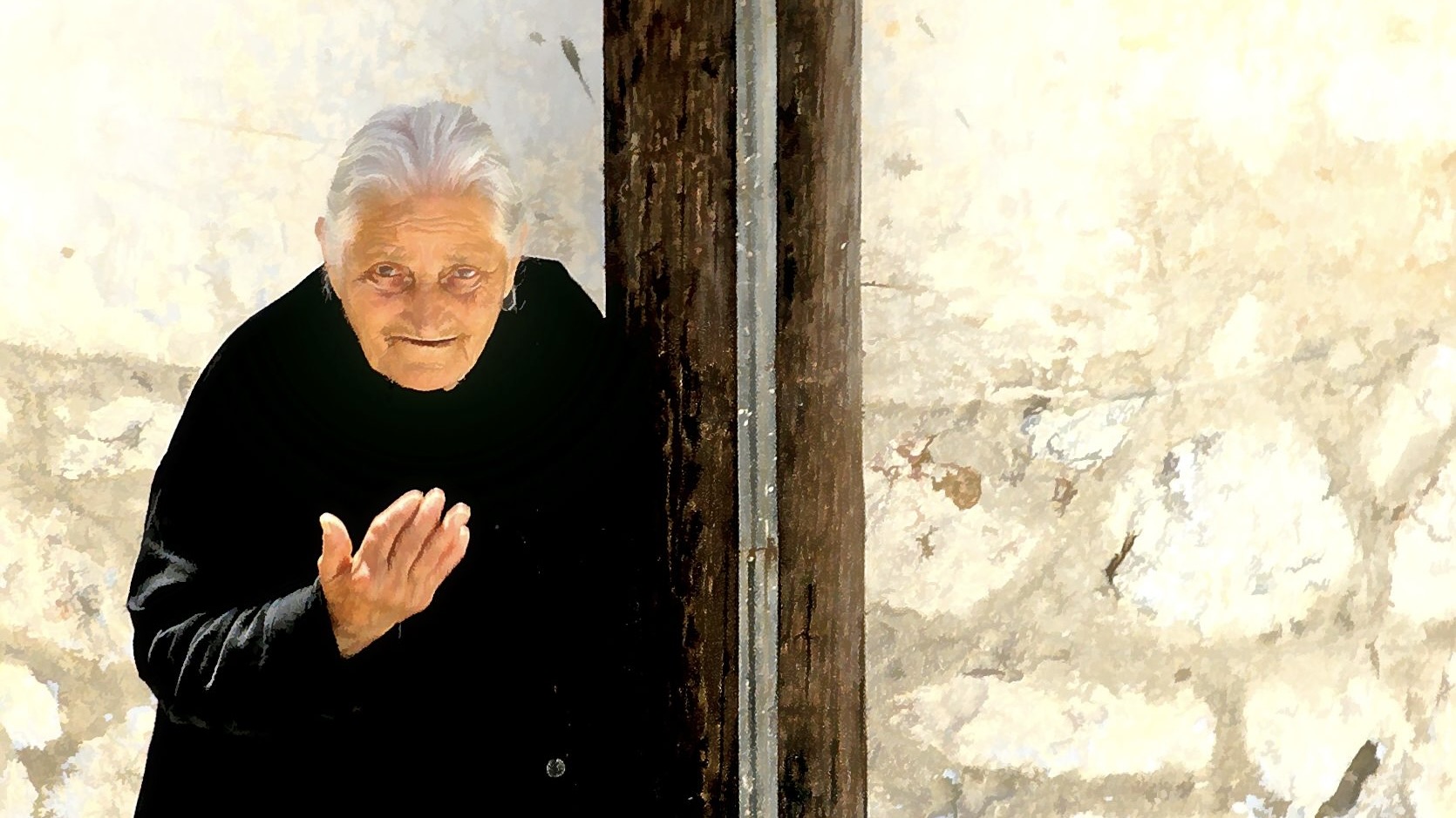 Old woman in black against a white stone wall.